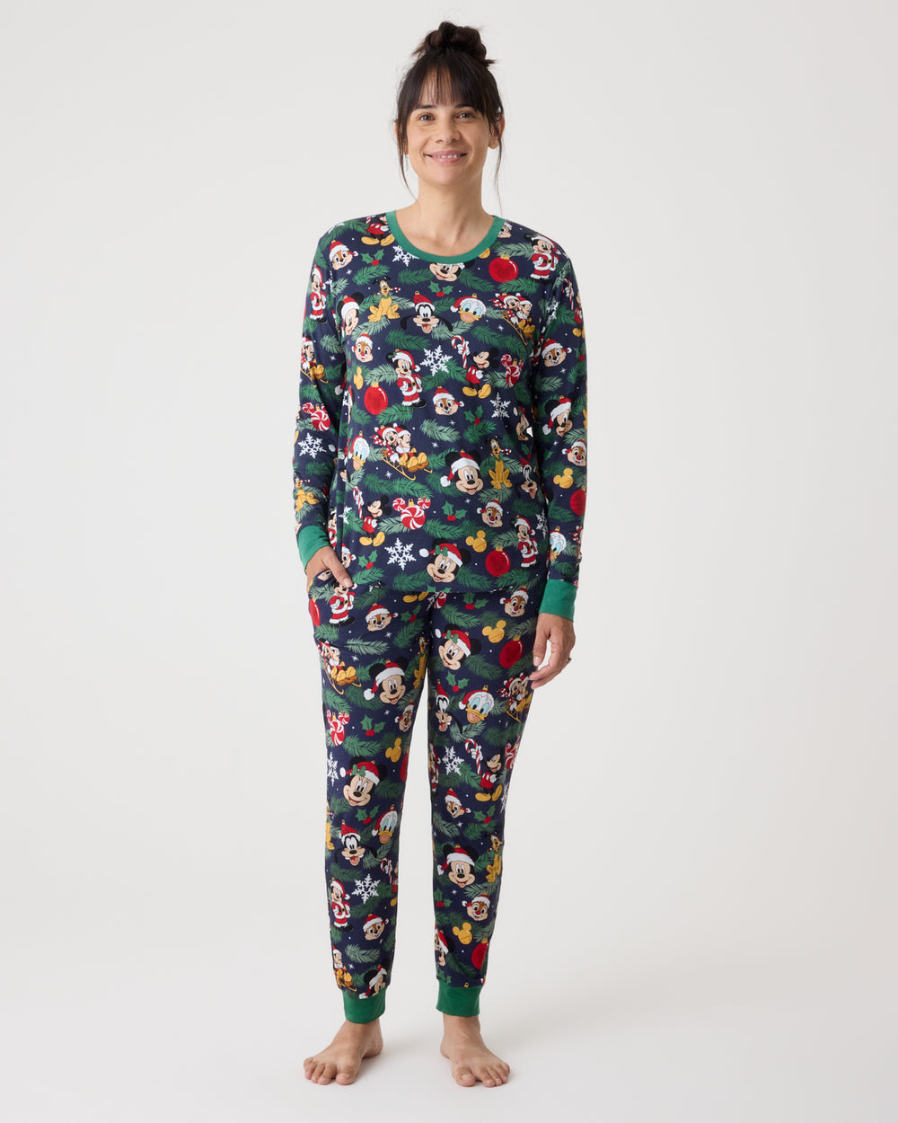 A woman stands barefoot against a plain light gray studio background wearing a long-sleeve pajama set with an allover Mickey Mouse and floral print. The pajamas are dark green with colorful cartoon and flower motifs, plus solid green cuffs and collar.