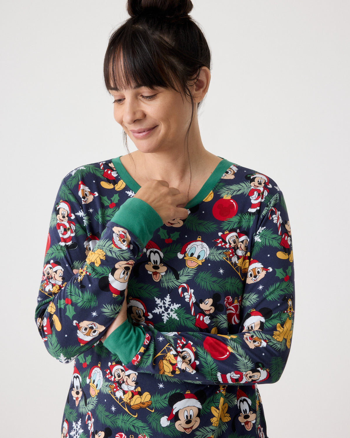 A woman wearing a long-sleeve Christmas-themed pajama top with a navy blue background and an all-over print of Disney characters like Mickey Mouse, Donald Duck, and Goofy, along with green cuffs and collar.