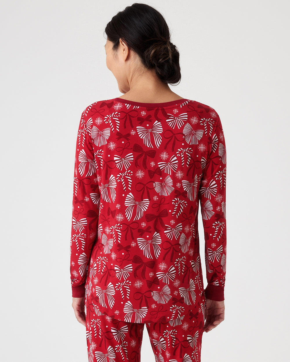 A woman viewed from the back wearing a red long-sleeve pajama top and matching pants with a white holiday-themed pattern of bows and snowflakes, against a light studio background.