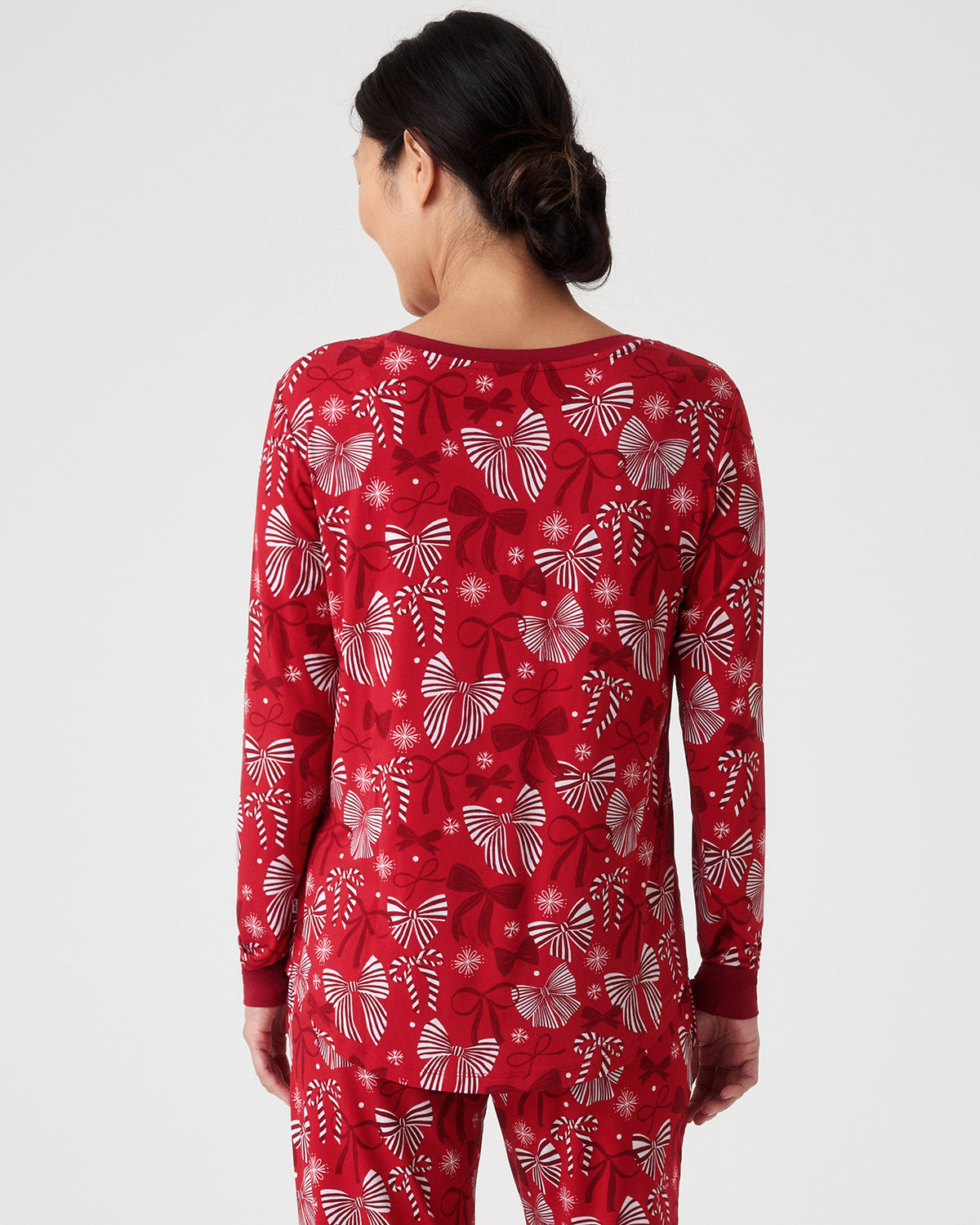 A woman viewed from the back wearing a red long-sleeve pajama top and matching pants with a white holiday-themed pattern of bows and snowflakes, against a light studio background.