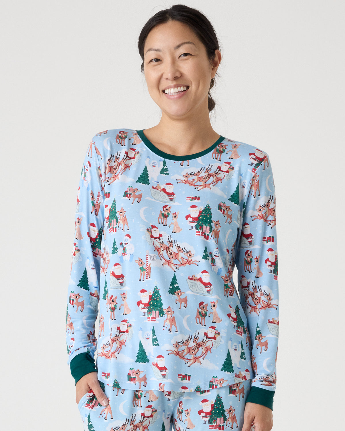 A woman smiling while wearing a light blue long-sleeve pajama top with a Christmas print of Santa Claus, reindeer, trees, and gifts, paired with matching pajama pants against a light gray studio background.