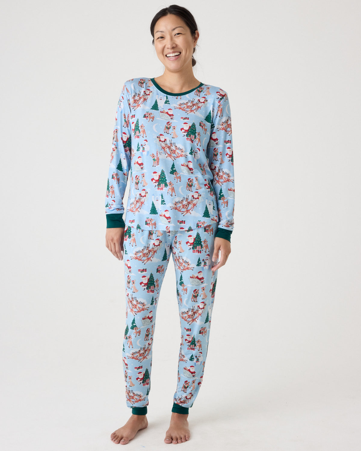 A woman modeling a light blue long-sleeve pajama set with a Christmas-themed print of Santa Claus, reindeer, snowman, and pine trees, against a plain white studio background.