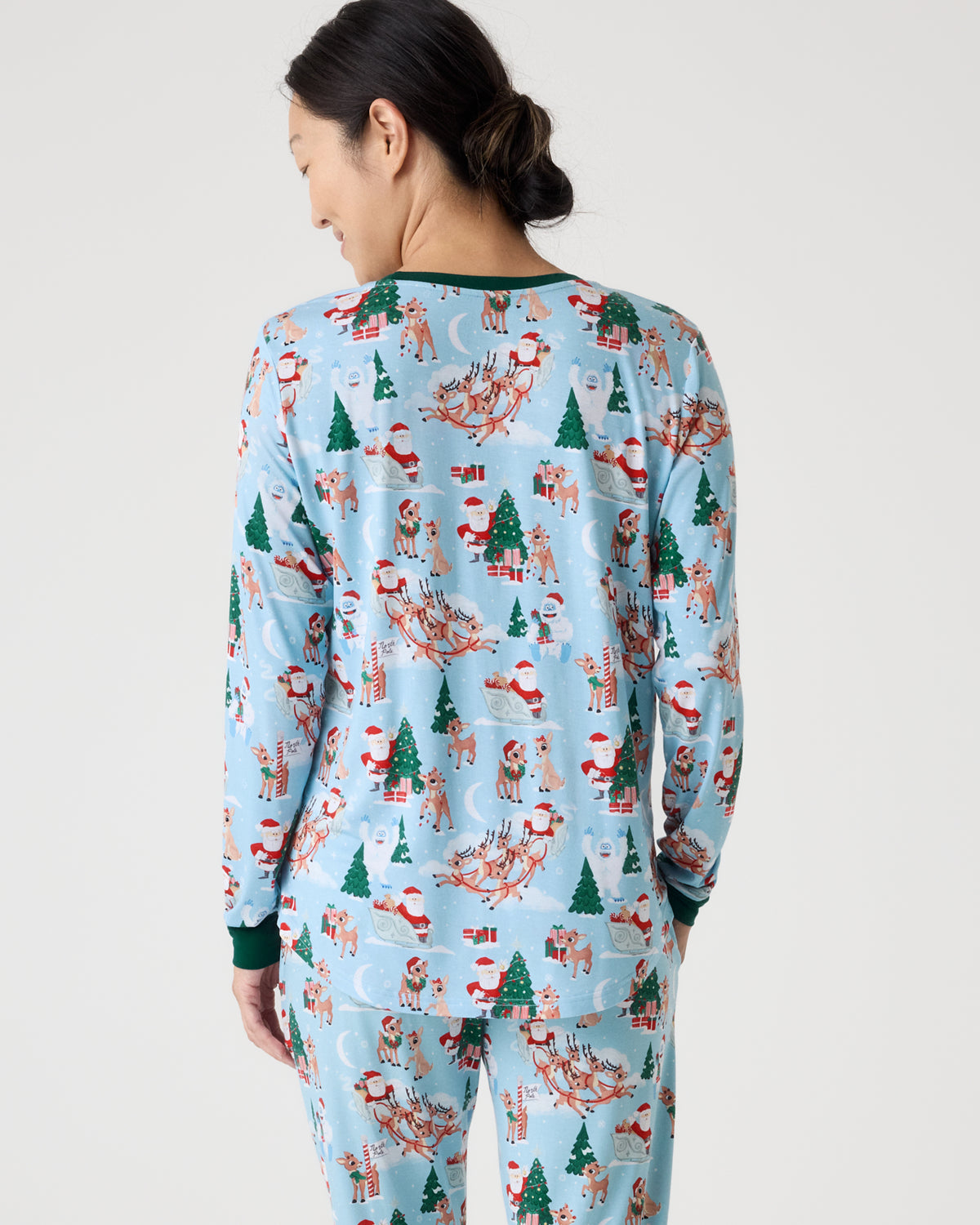 A woman shown from the back wearing a light blue long-sleeve pajama set with a festive Christmas print featuring Santa Claus, reindeer, Christmas trees, and sleighs, against a plain light gray studio background.