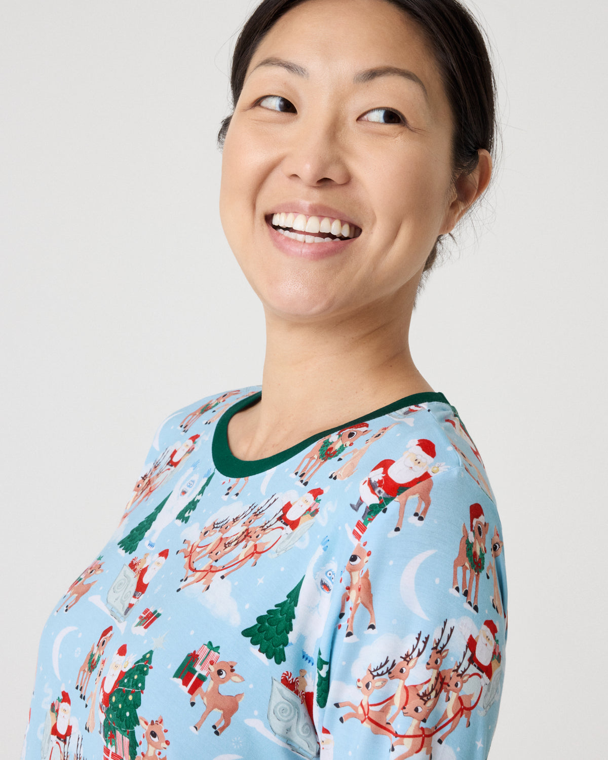 A woman smiling and looking to the side, wearing a light blue long-sleeve pajama top with a green crew neck and a festive Christmas print featuring Santa Claus, reindeer, and Christmas trees.