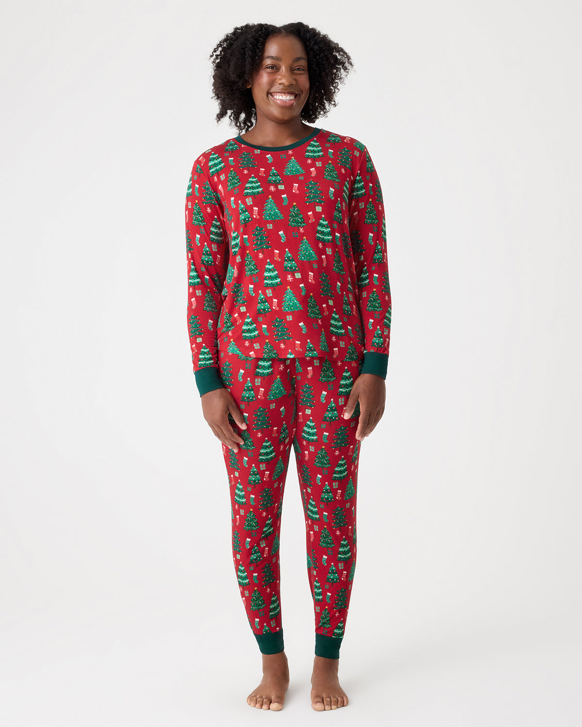 A woman modeling a red long-sleeve pajama set with a green Christmas tree pattern, standing against a plain light gray studio background.