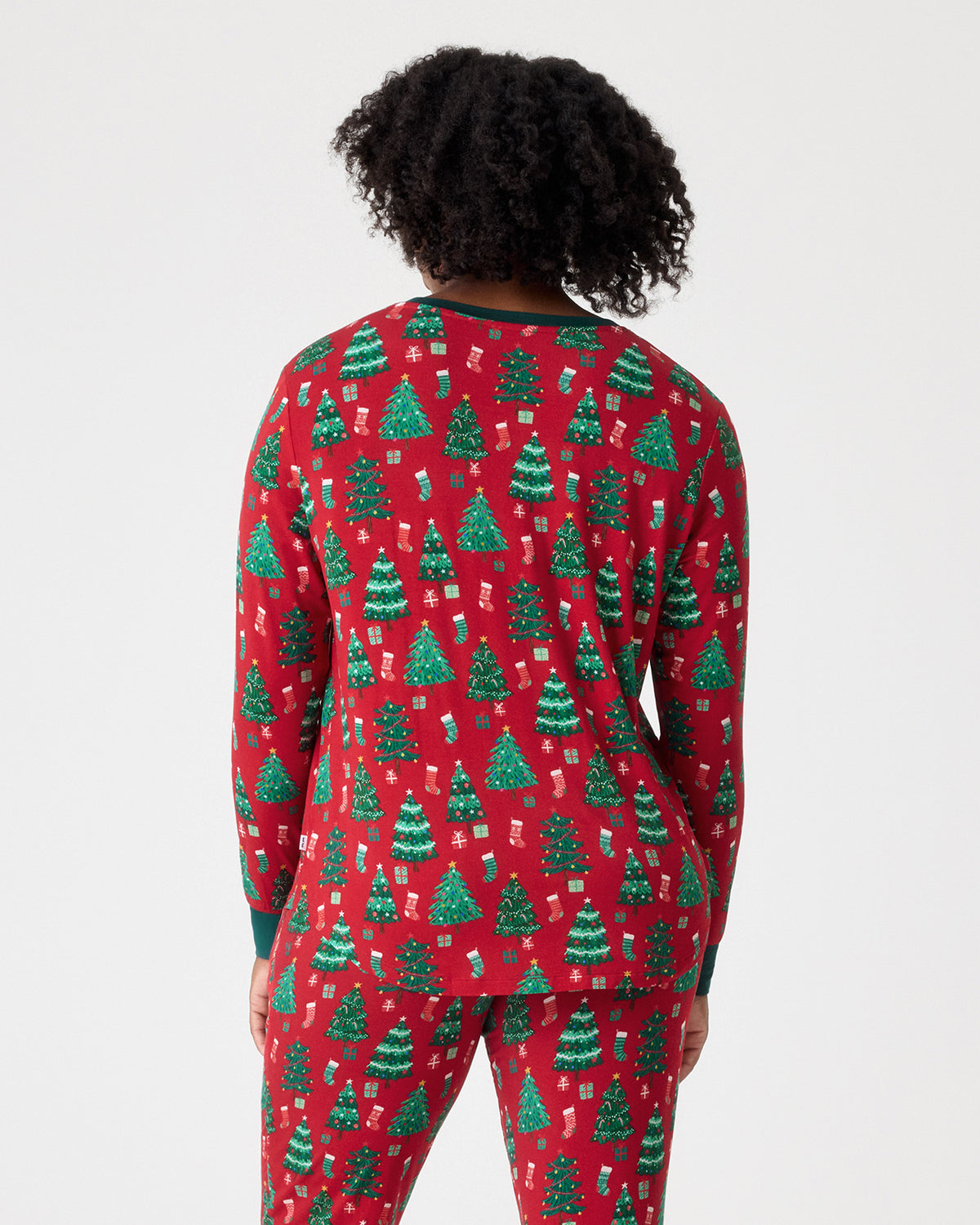 A person shown from the back wearing a red long-sleeve pajama top and matching pants with an allover green Christmas tree print, against a light gray studio background.