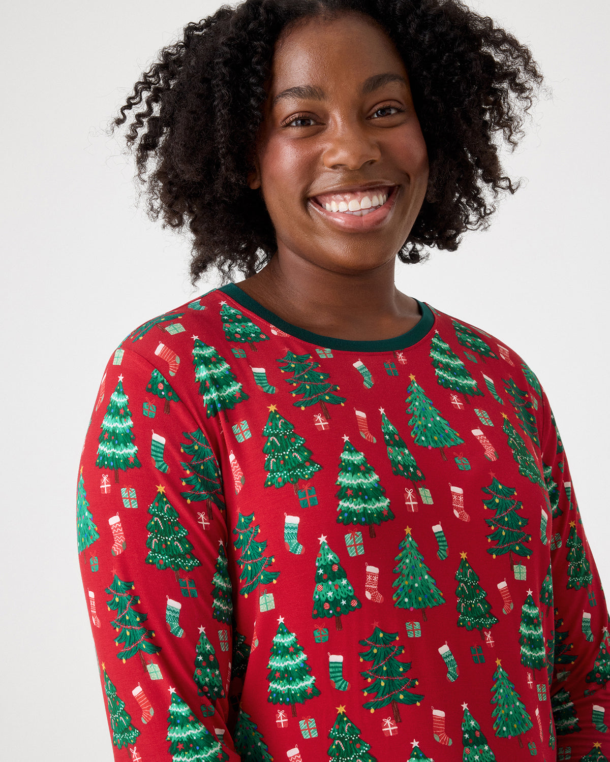 A smiling woman wearing a red long-sleeve pajama top with a green Christmas tree and stocking pattern, photographed against a light gray studio background.
