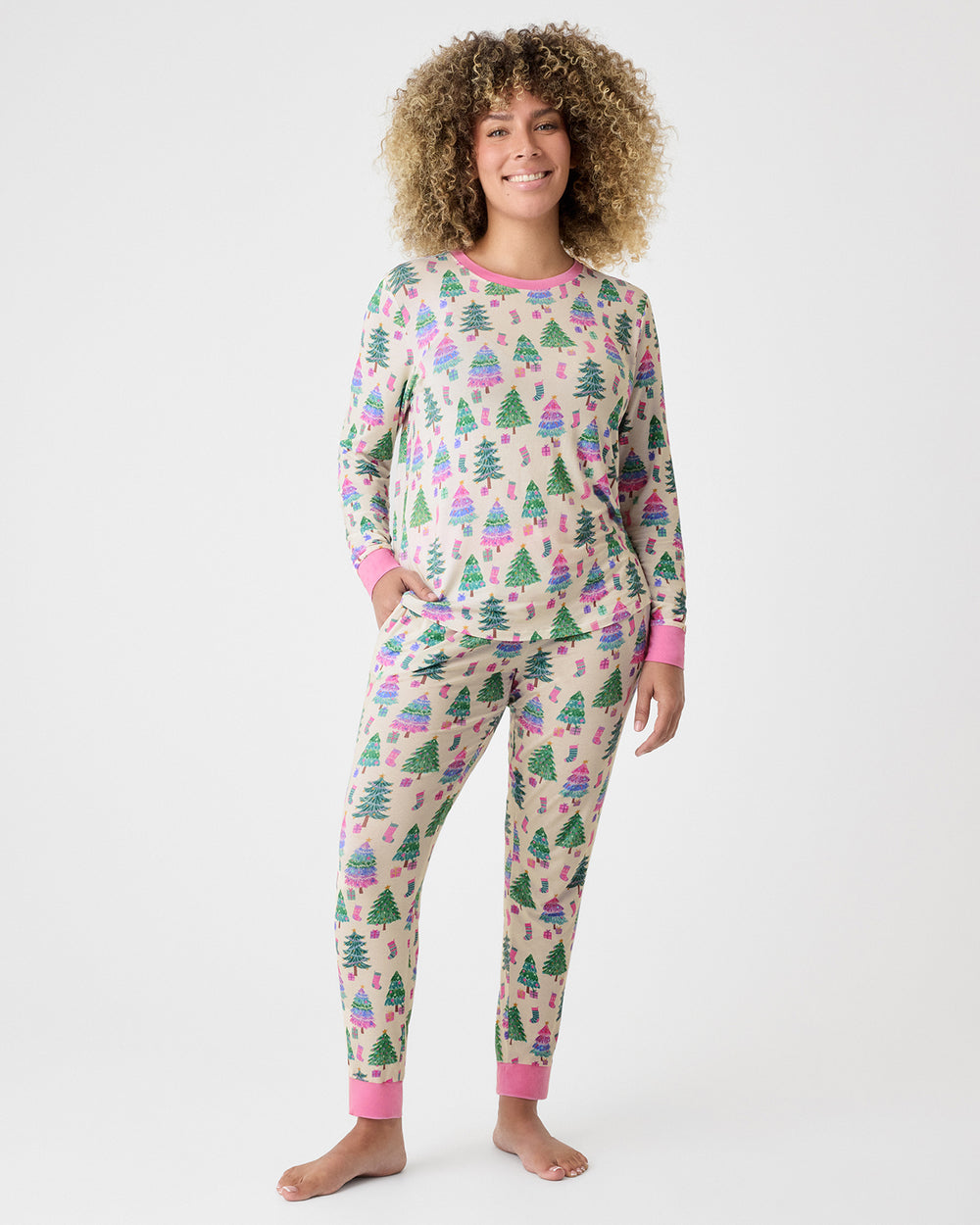 A woman modeling a two-piece long-sleeve pajama set with a Christmas tree print on a beige background with pink cuffs and collar.