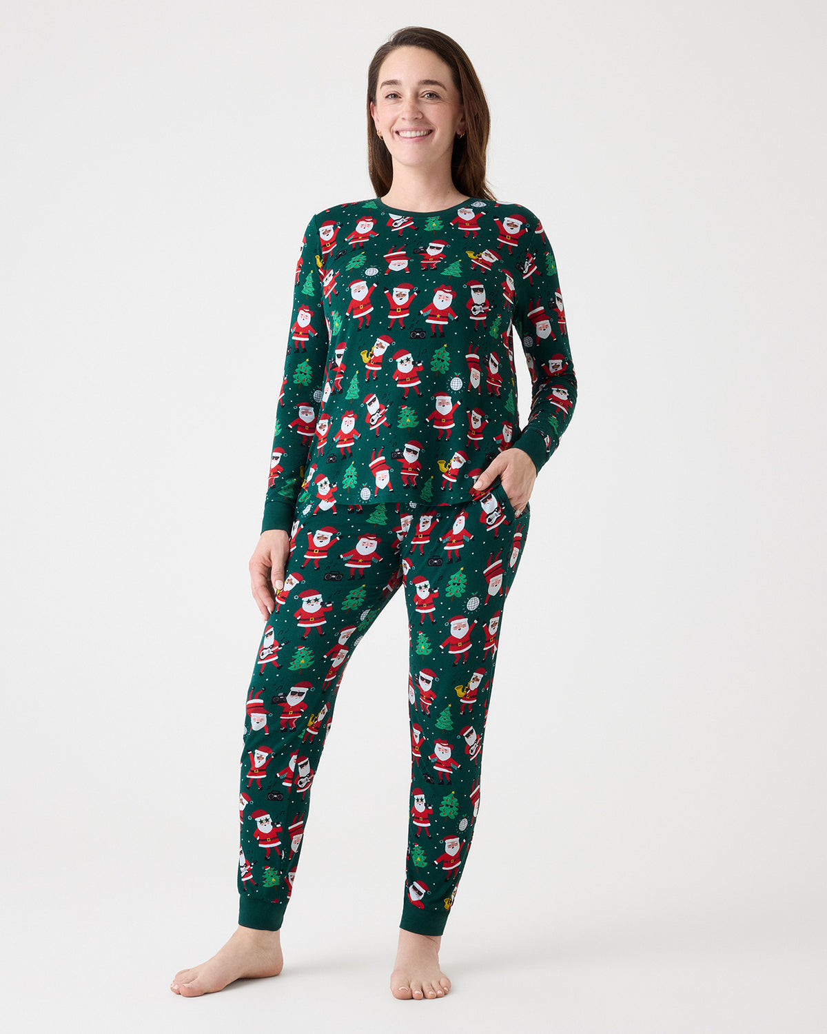 A woman modeling a dark green long-sleeve pajama set with an all-over Santa Claus and Christmas tree pattern, standing against a plain light gray studio background.
