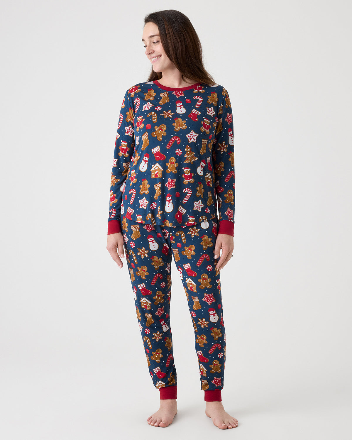 A woman modeling a long-sleeve pajama set with a festive Christmas print on a solid light gray background. The pajamas feature a navy blue base with a colorful pattern of gingerbread cookies, candy canes, snowmen, and stockings, finished with solid red cuffs and collar.
