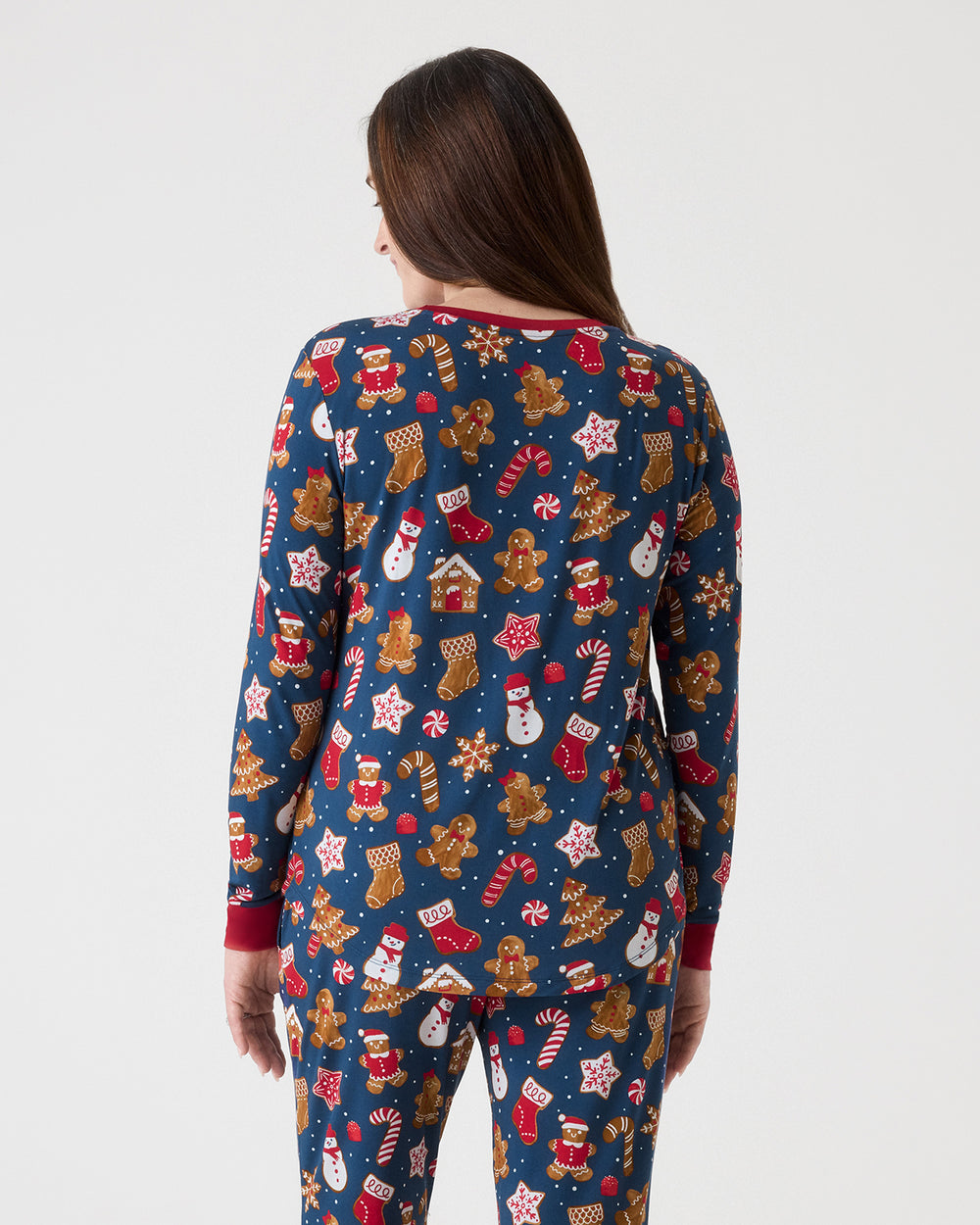 A woman viewed from the back wearing a navy blue long-sleeve pajama set with a festive Christmas print featuring gingerbread men, Santa Claus, snowmen, candy canes, and stars. The pajamas have red cuffs and a red collar, and are photographed against a solid light gray background.