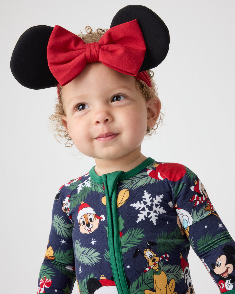 A toddler wearing a festive holiday zip-up one-piece pajama with a green collar and a colorful cartoon print, accessorized with a black and red Minnie Mouse-inspired headband, photographed against a light gray studio background.
