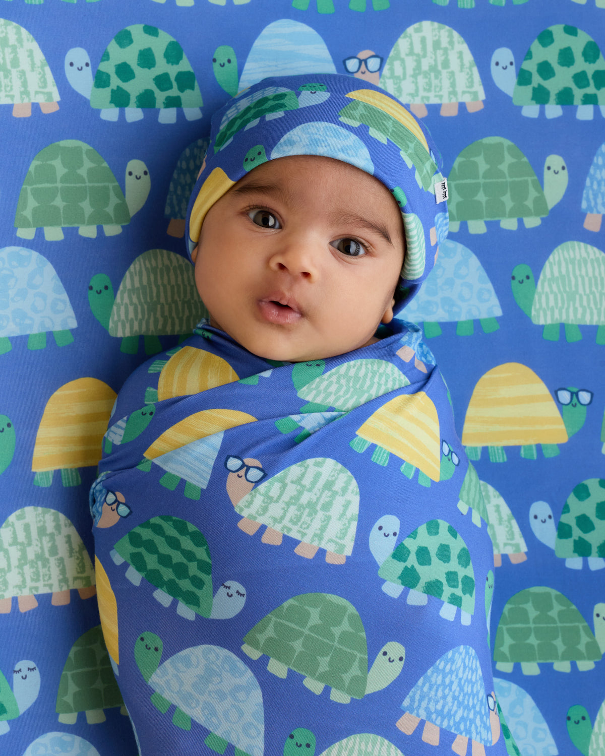 Newborn wrapped in the Shell Parade Swaddle & Hat Set on a matching crib sheet