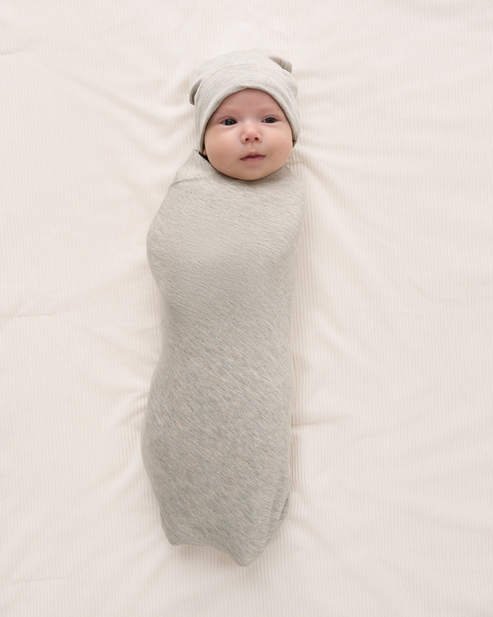 A baby swaddled in a light gray textured wrap and wearing a matching gray beanie, lying on a cream-colored fabric background.