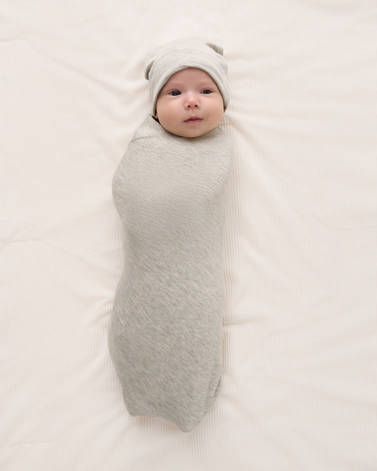 A baby swaddled in a light gray textured wrap and wearing a matching gray beanie, lying on a cream-colored fabric background.