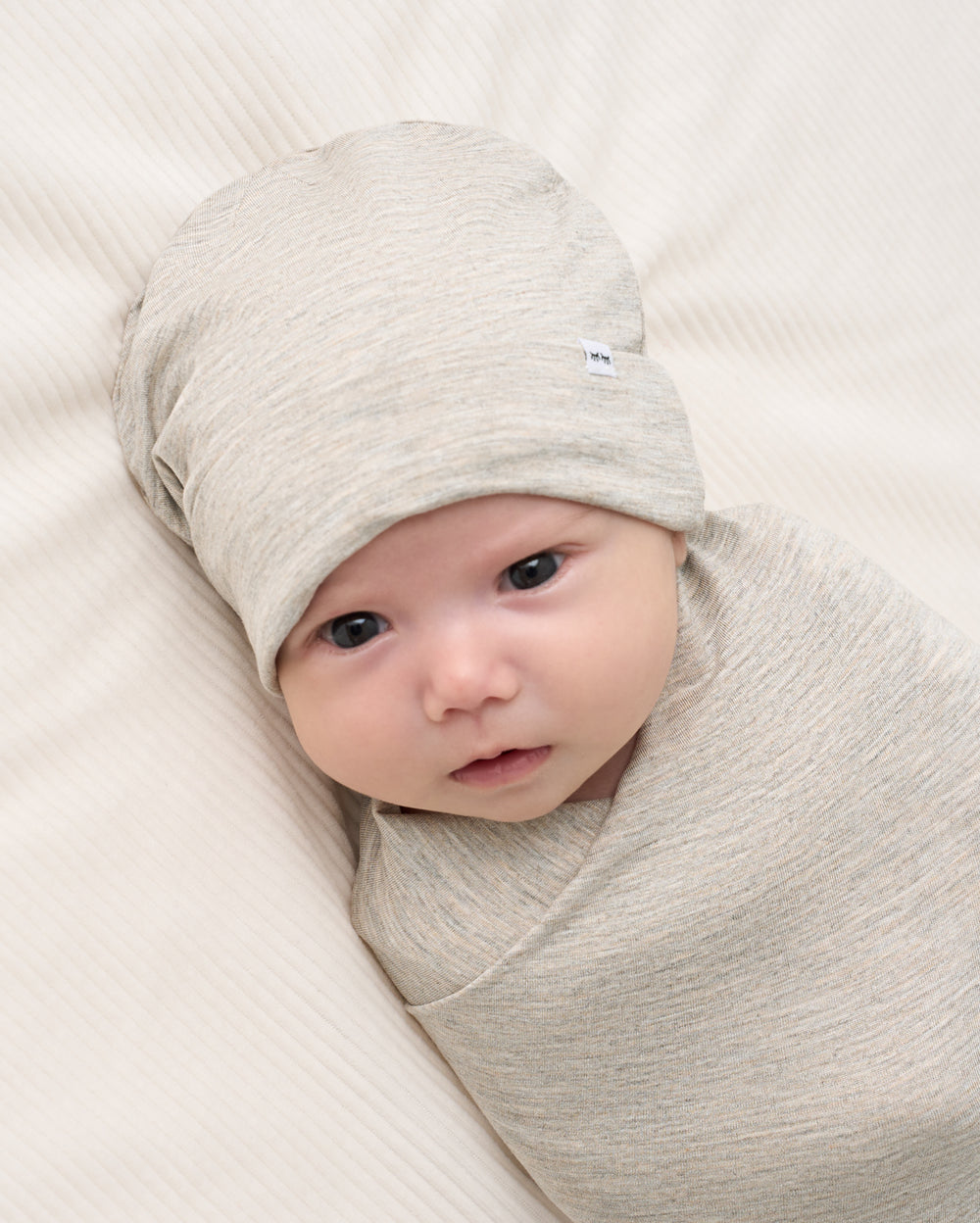 A baby swaddled in a light heather gray wrap blanket and wearing a matching knit beanie, lying on a textured cream-colored blanket.