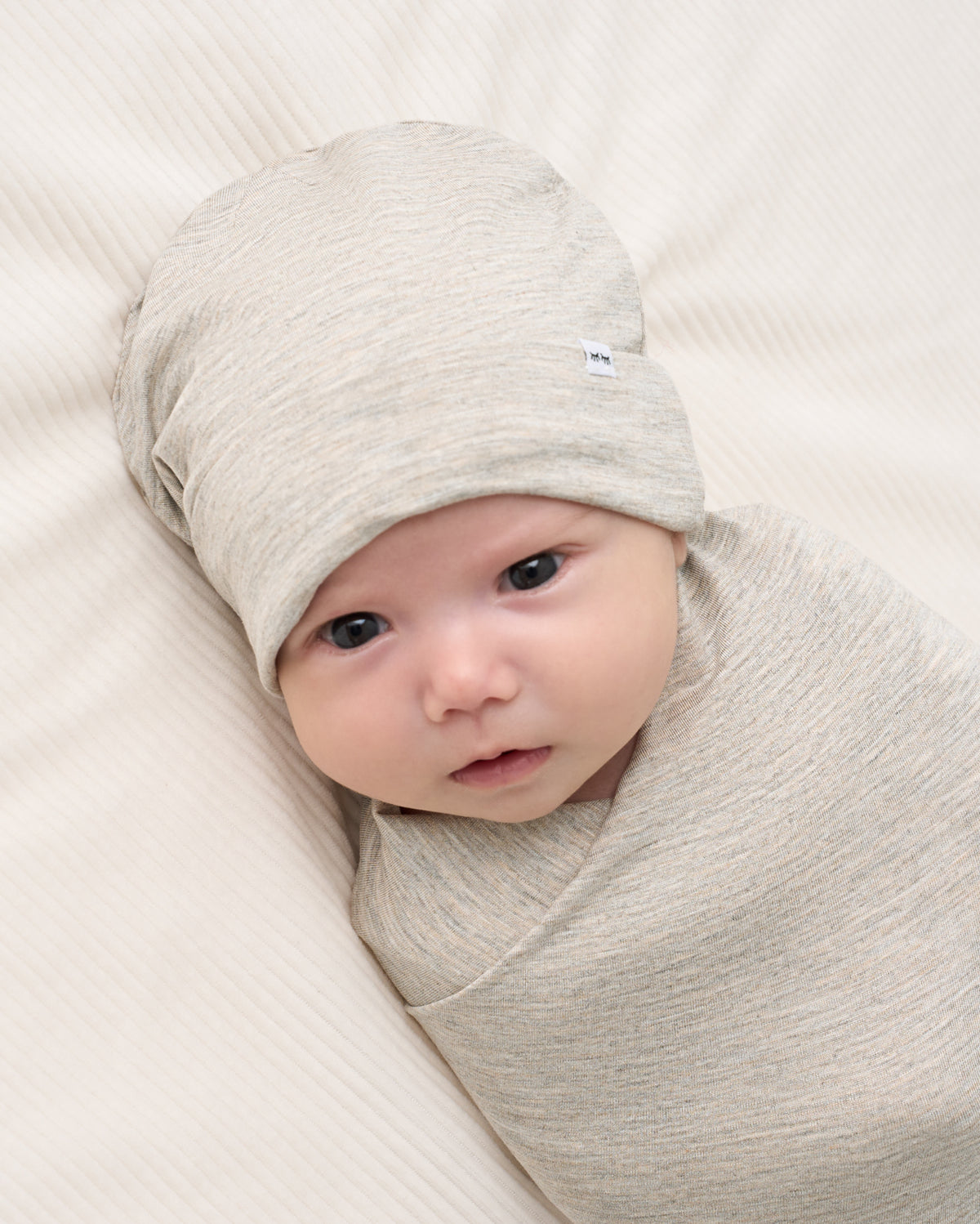 A baby swaddled in a light heather gray wrap blanket and wearing a matching knit beanie, lying on a textured cream-colored blanket.