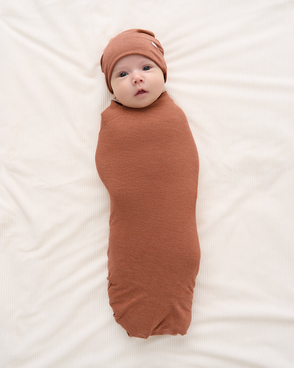 A baby swaddled in a rust-brown fabric wrap and matching beanie hat, lying on a white textured blanket.