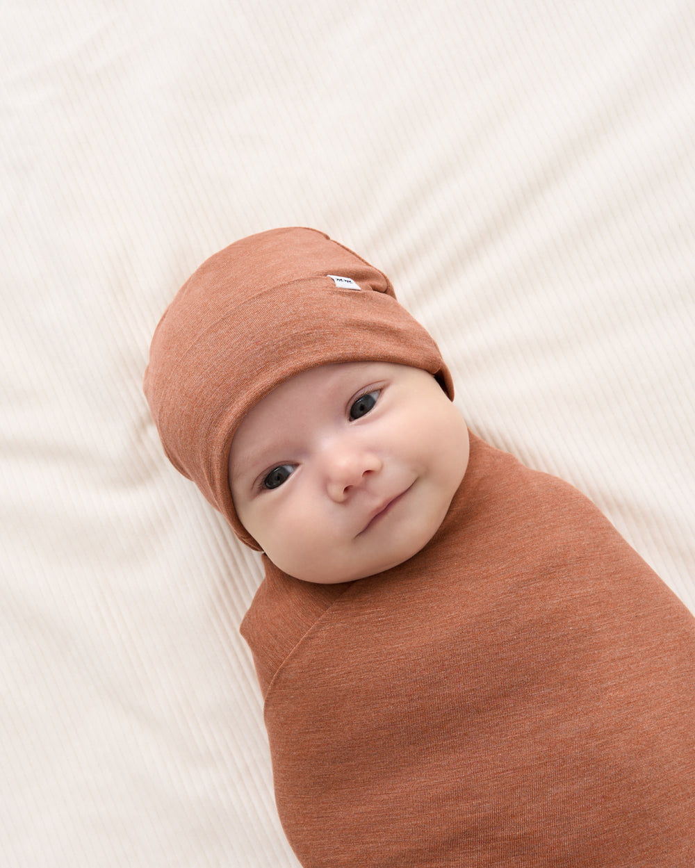 A baby swaddled in a rust-colored blanket and wearing a matching knit bonnet, lying on a textured cream blanket.