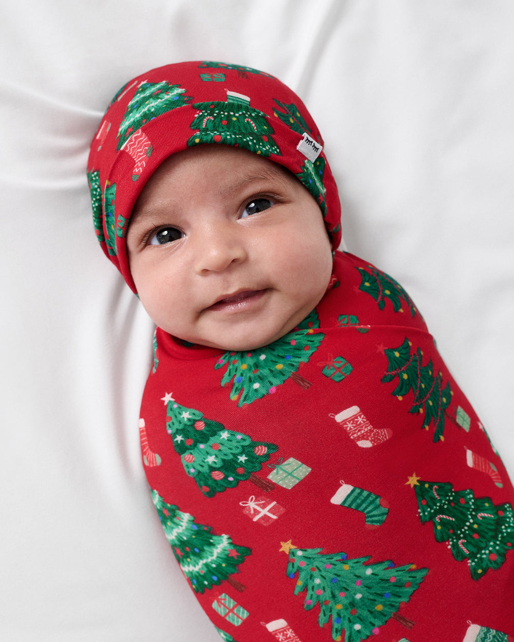 A baby swaddled in a red holiday-themed swaddle blanket and matching beanie, featuring a green Christmas tree and stocking pattern, lying on a white fabric background.
