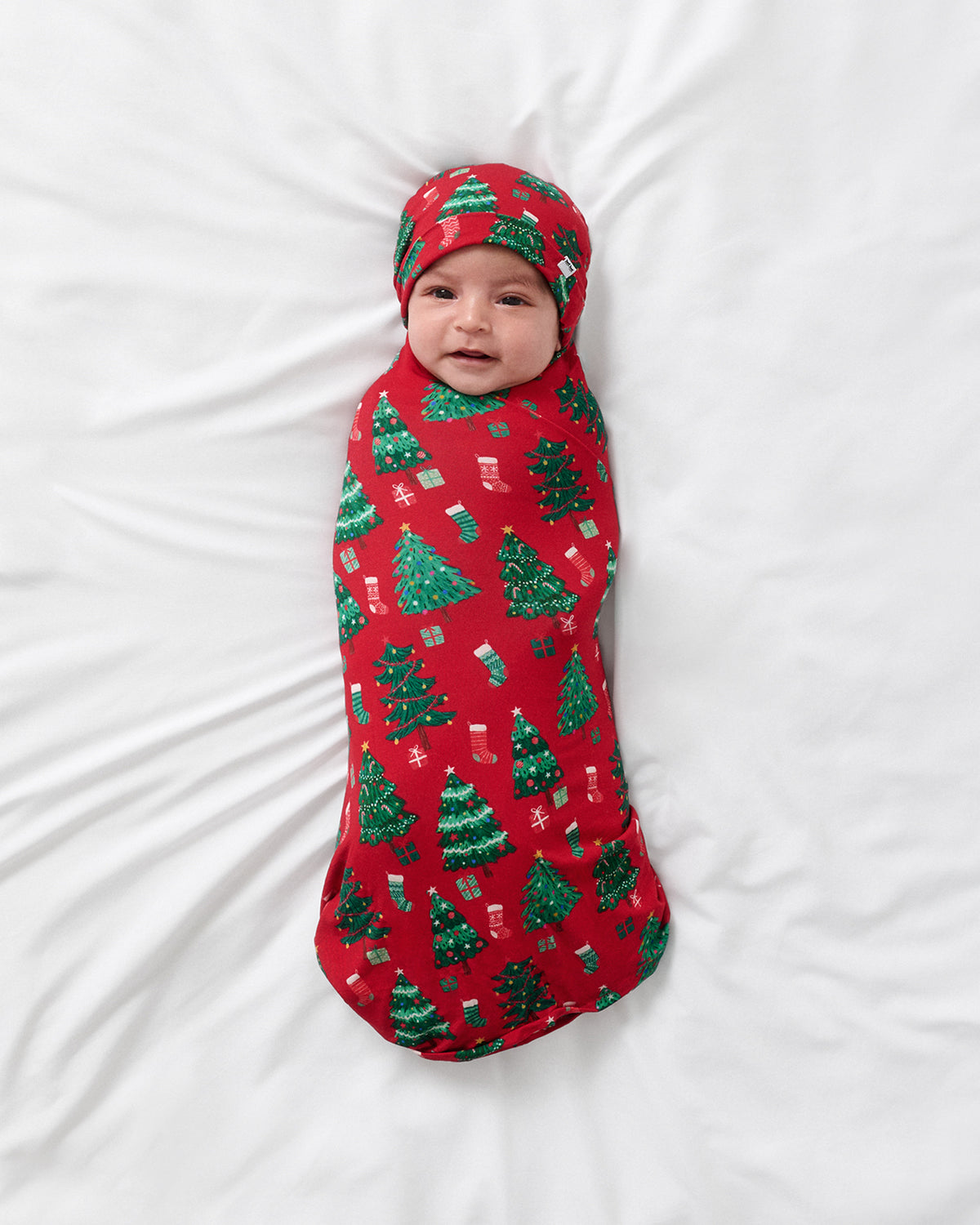 A baby swaddled in a red Christmas-themed swaddle blanket with a matching red hat, lying on white bedding. The blanket features a pattern of green sequined Christmas trees, gifts, and stockings.
