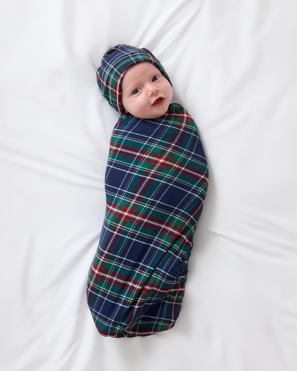 A baby swaddled in a navy blue tartan plaid blanket with matching beanie hat, lying on white sheets.