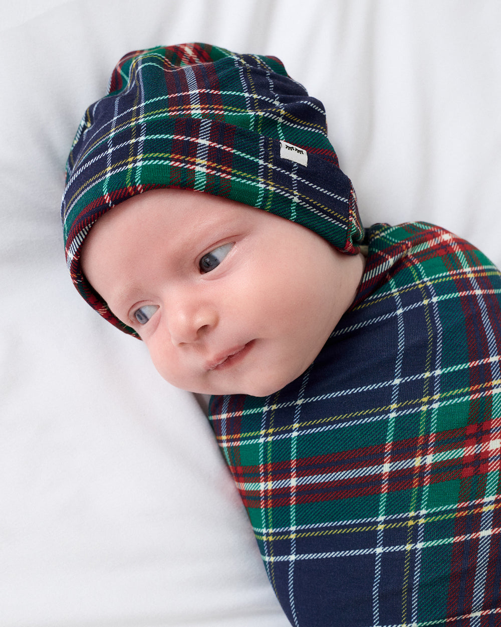 A baby wearing a navy blue beanie and swaddled in a matching green, red, and navy tartan plaid blanket, lying on a white blanket.