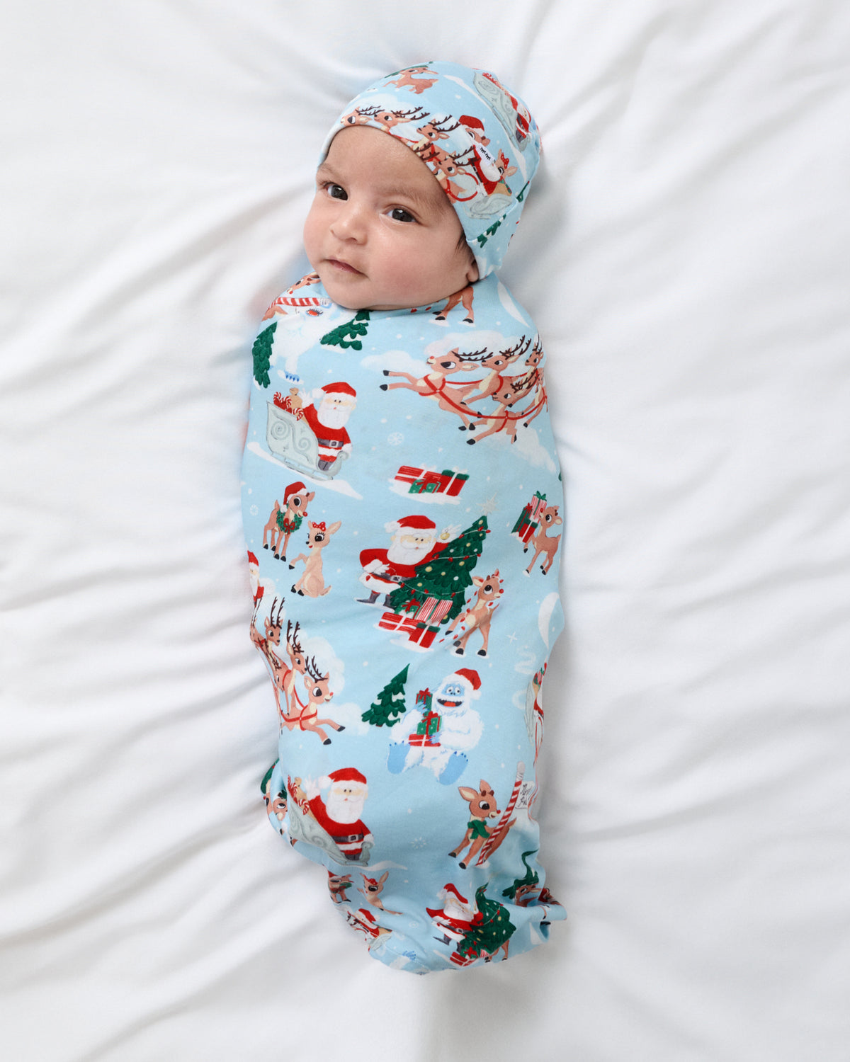 A baby swaddled in a light blue Christmas-themed blanket with a matching bonnet, lying on white bedding. The blanket features a festive print of Santa Claus, reindeer, snowmen, and Christmas trees.