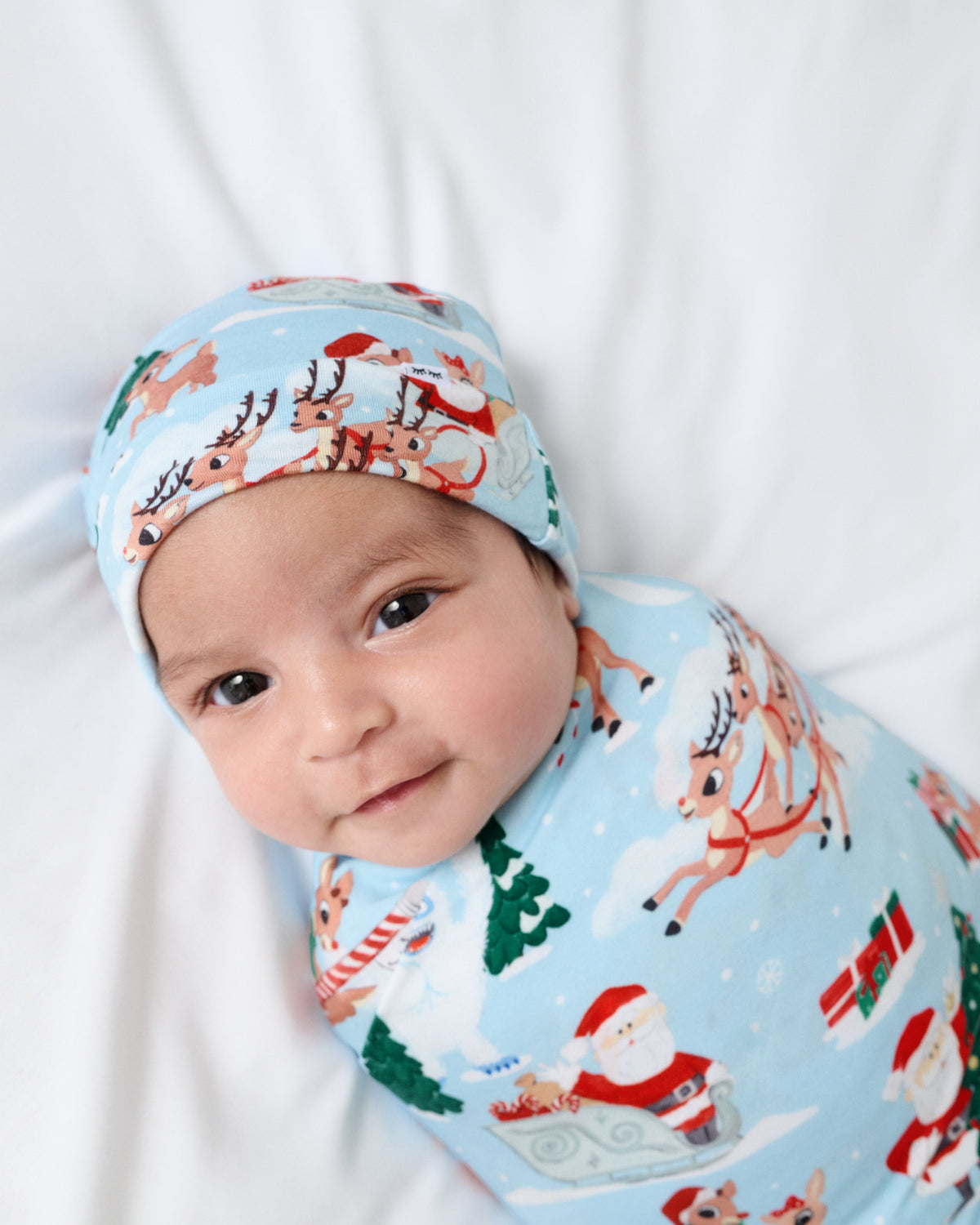 A baby lying on a white blanket, wearing a light blue holiday-themed swaddle wrap and matching beanie hat with a print of Santa Claus, reindeer, and Christmas trees.