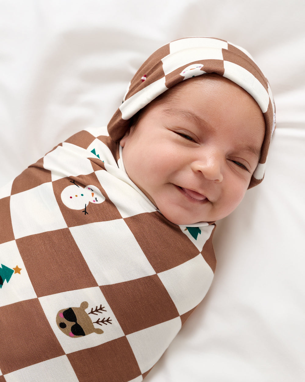 A smiling newborn baby wrapped in a brown and cream checkered swaddle blanket with holiday-themed prints, wearing a matching checkered hat, lying on a white background.