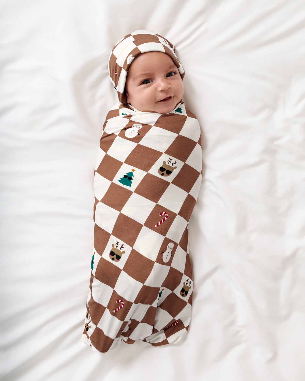 A baby swaddled in a brown and cream checkered blanket with small holiday motifs, wearing a matching beanie, lying on a white bed.