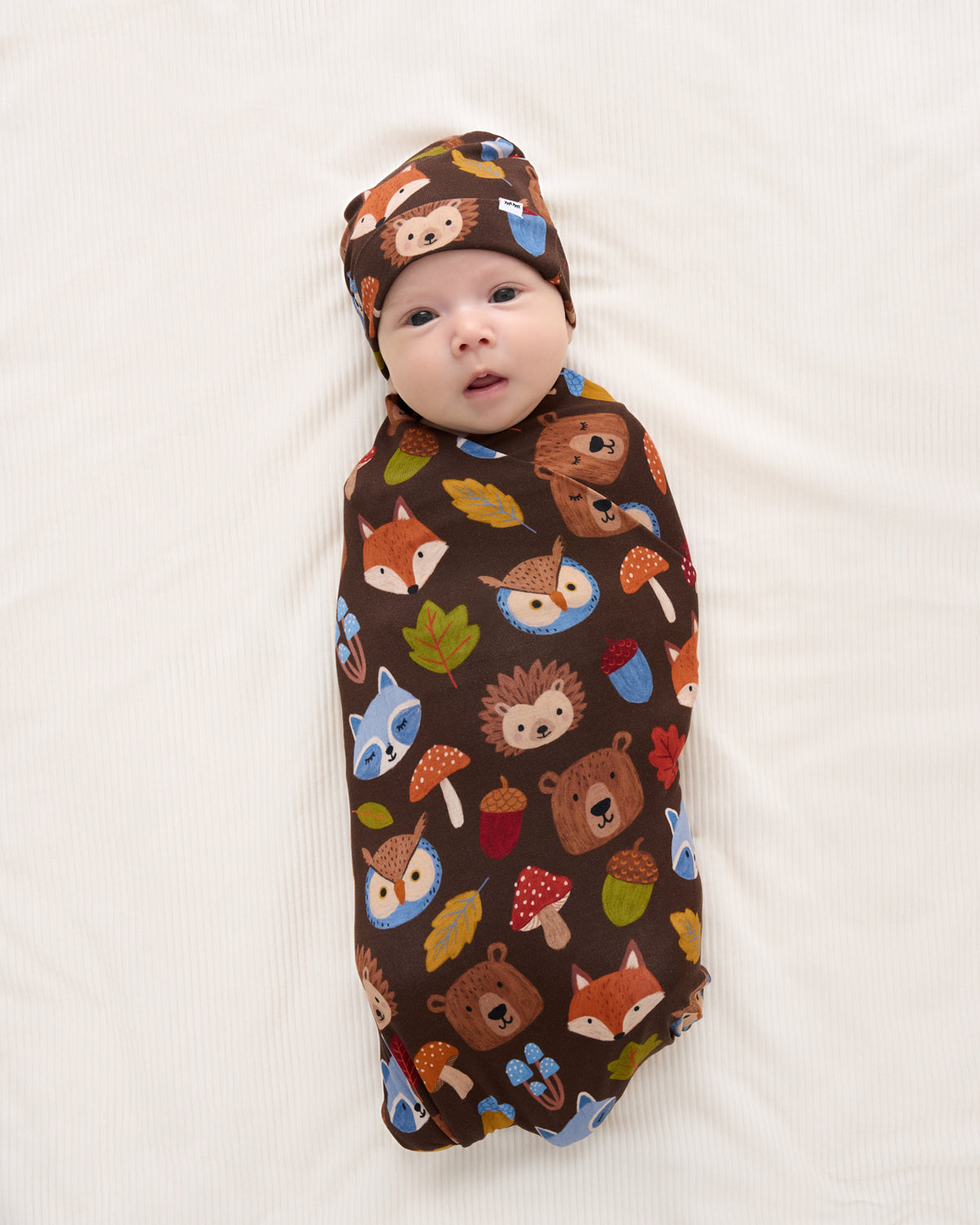 A baby swaddled in a dark brown fabric wrap with a colorful woodland animal print, including foxes, hedgehogs, bears, acorns, and leaves, along with a matching bonnet, lying on a white textured blanket.