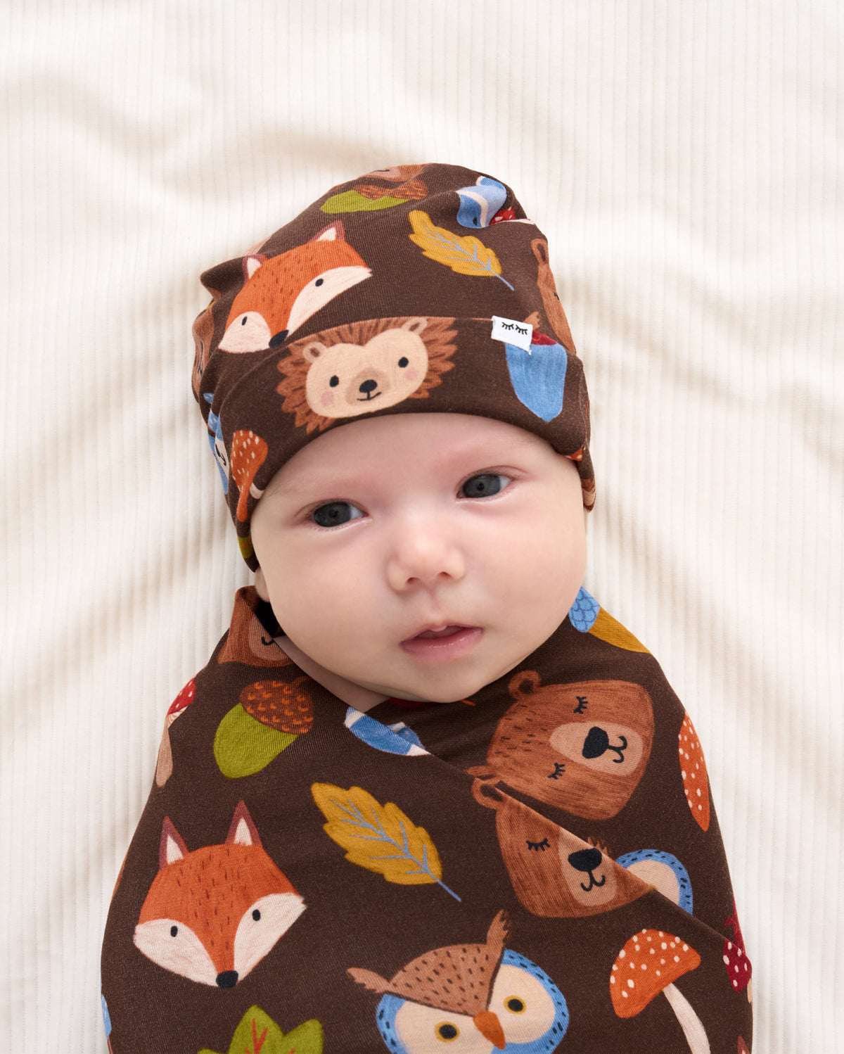 A baby swaddled and wearing a matching brown forest-themed swaddle wrap and bonnet with printed fox, hedgehog, bear, owl, acorn, and leaf motifs, photographed on a soft cream fabric background.
