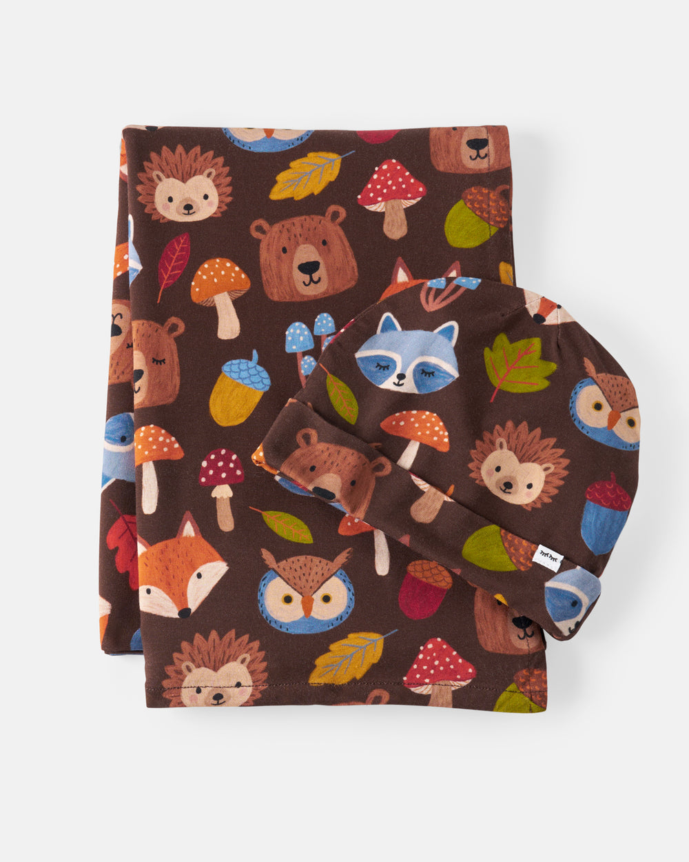 A folded fabric set with a woodland animal print on a dark brown background, including a rectangular cloth piece and a rounded hat or pouch, featuring illustrated bears, foxes, owls, mushrooms, acorns, and leaves.