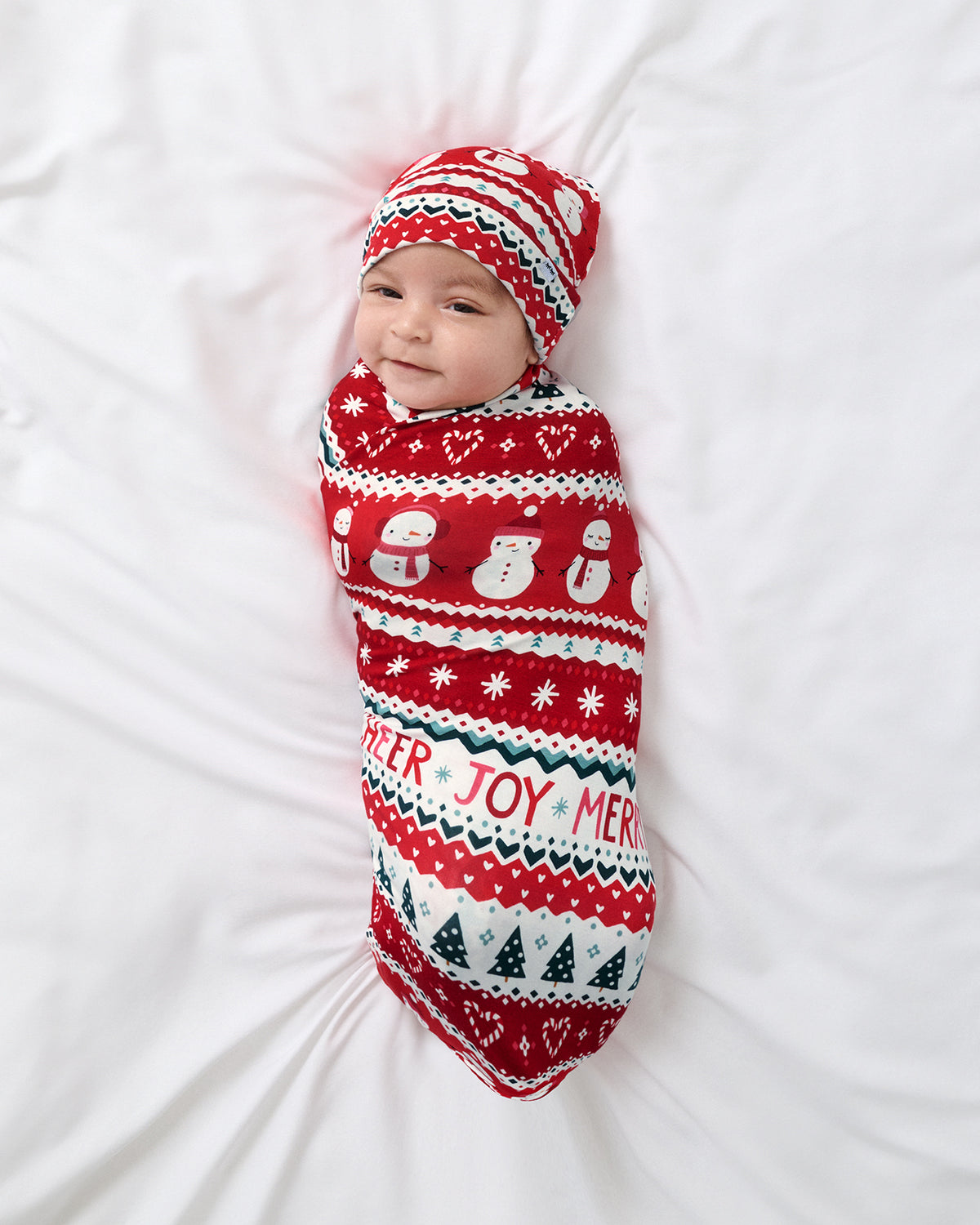 A baby swaddled in a red and white holiday knit blanket with a matching beanie, lying on a white bed. The knit pattern features snowmen, snowflakes, Christmas trees, and the words 'Cheer' 'Joy' and 'Merry'.