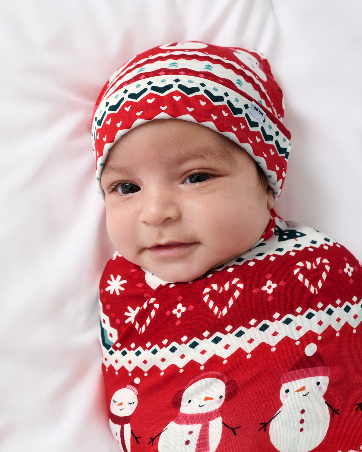 A baby swaddled in a red and white Christmas-themed blanket with snowman prints and wearing a matching knit hat, lying on a white background.