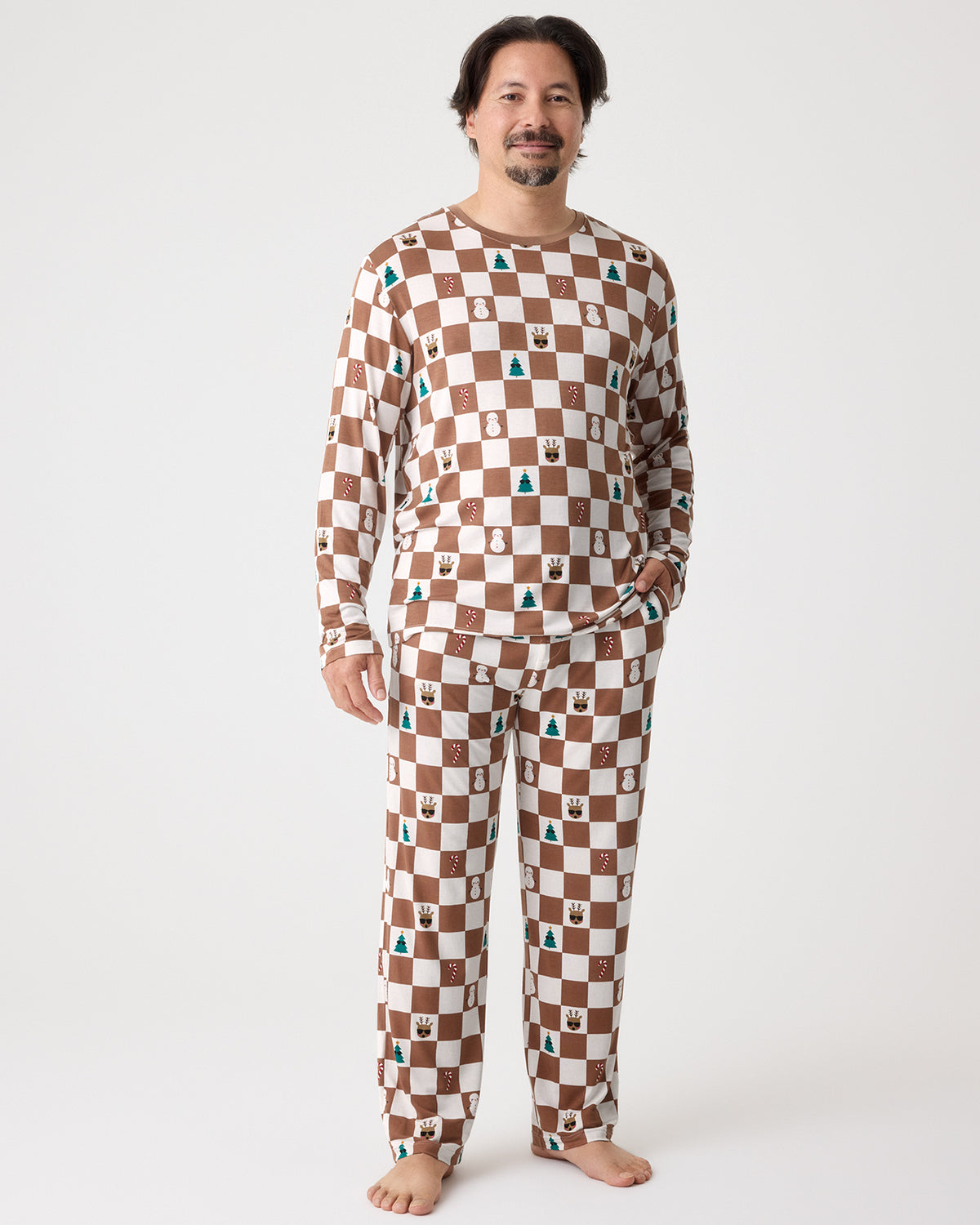 A man wearing a matching long-sleeve pajama set with a brown and cream checkerboard pattern and small Christmas icons, standing against a plain light gray studio background.