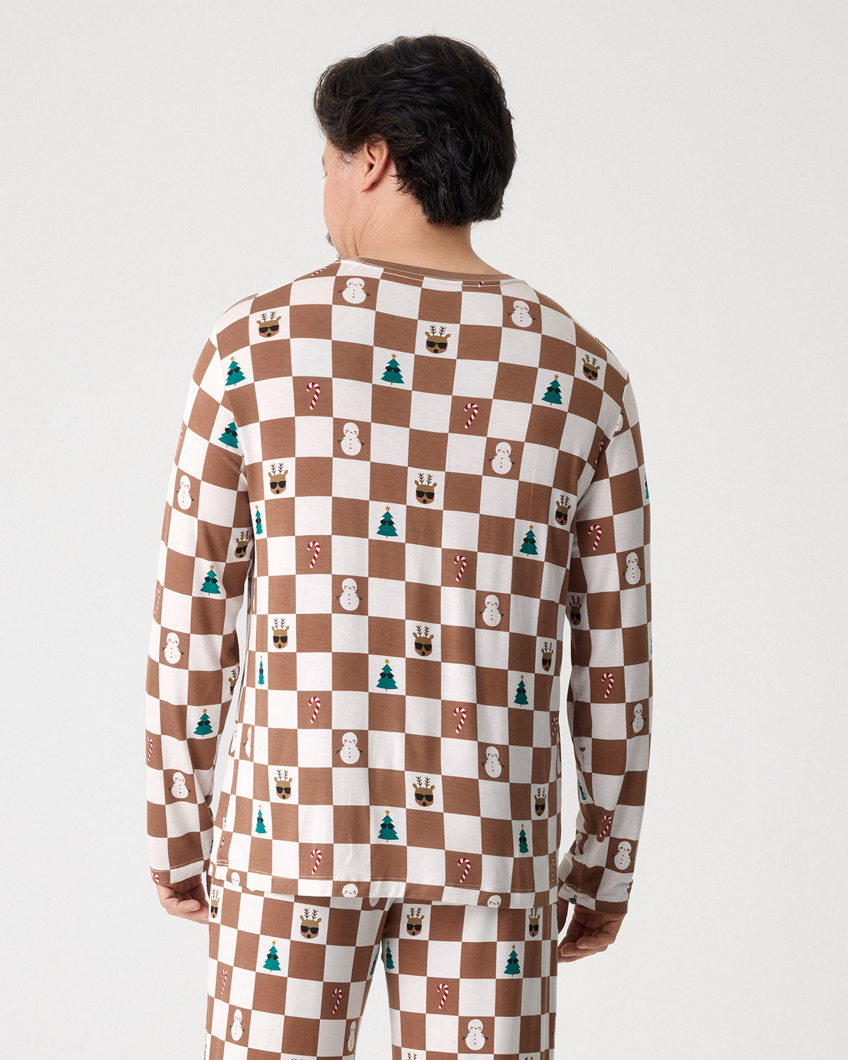 A man viewed from behind wearing a long-sleeve pajama top and matching pants with a brown and white checkerboard pattern decorated with small holiday motifs like snowmen, pine trees, and gingerbread houses, against a light gray studio background.