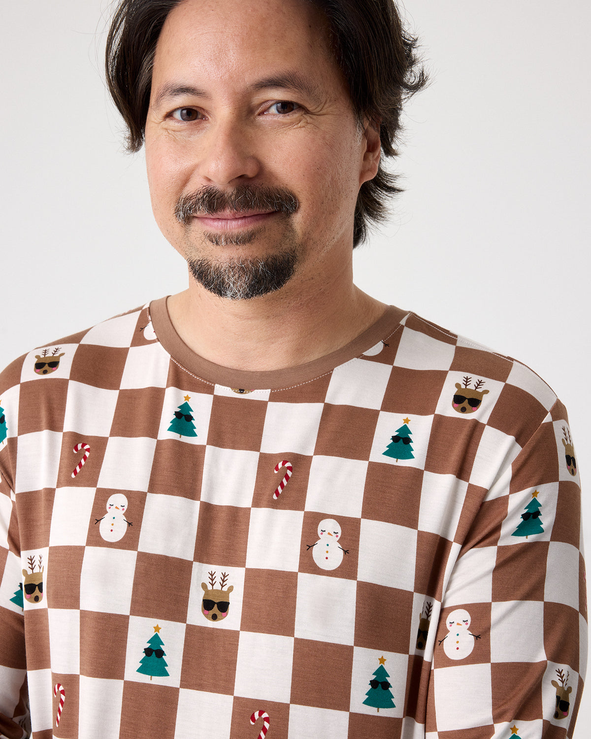 A man wearing a brown and white checkered long-sleeve shirt with a Christmas theme, featuring printed snowmen, Christmas trees, candy canes, and reindeer, against a plain light gray background.