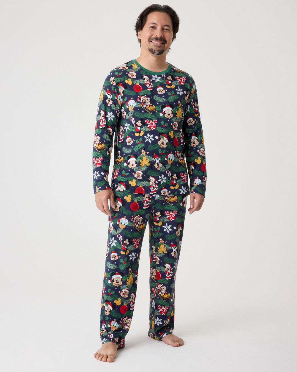 A man wearing a long-sleeve pajama set with a colorful Mickey Mouse and friends Christmas print, standing against a plain light gray studio background.