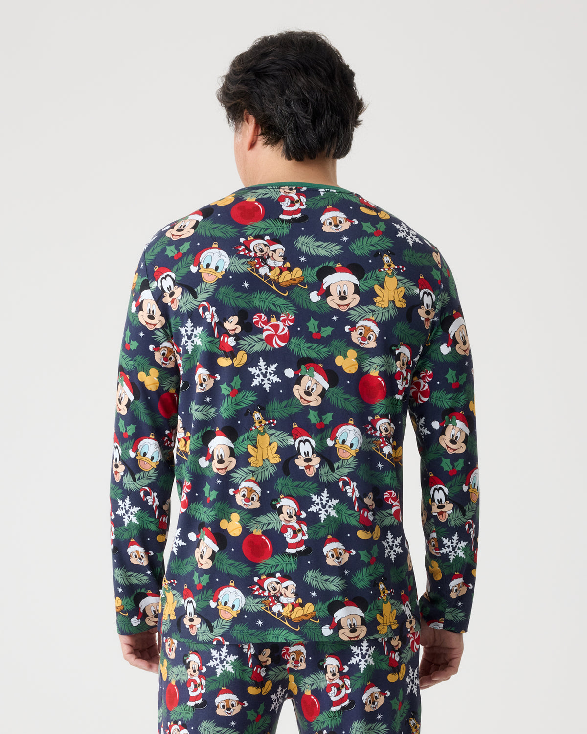 A man viewed from behind wearing a long-sleeve navy blue pajama set with an all-over festive Mickey Mouse and Friends print, set against a plain light gray background.