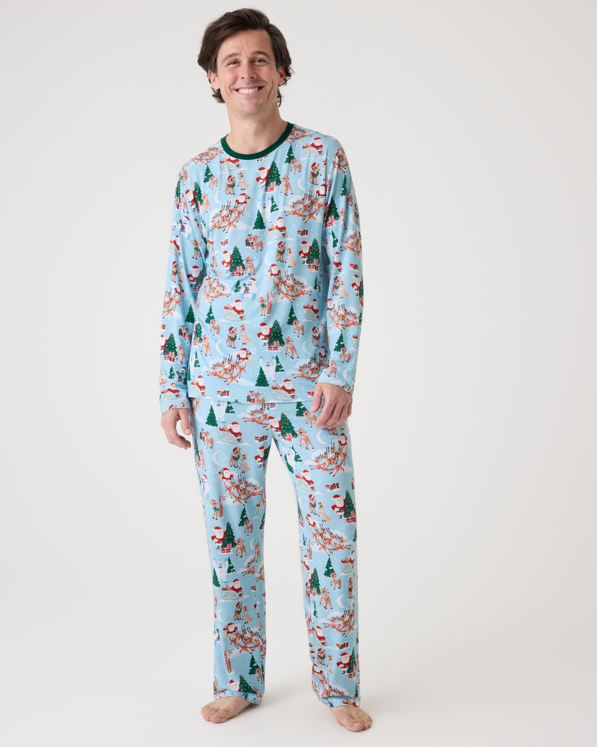 A man modeling a light blue long-sleeve pajama set with a Christmas tree and Santa pattern, standing against a plain white studio background.
