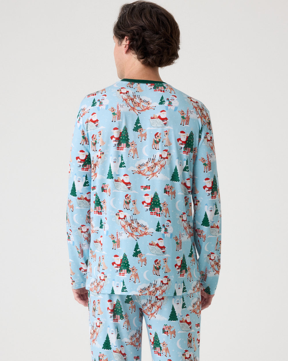 A person shown from the back wearing a light blue long-sleeve pajama set with an all-over Christmas print featuring Santa Claus, reindeer, Christmas trees, and snowman motifs, against a plain light gray studio background.