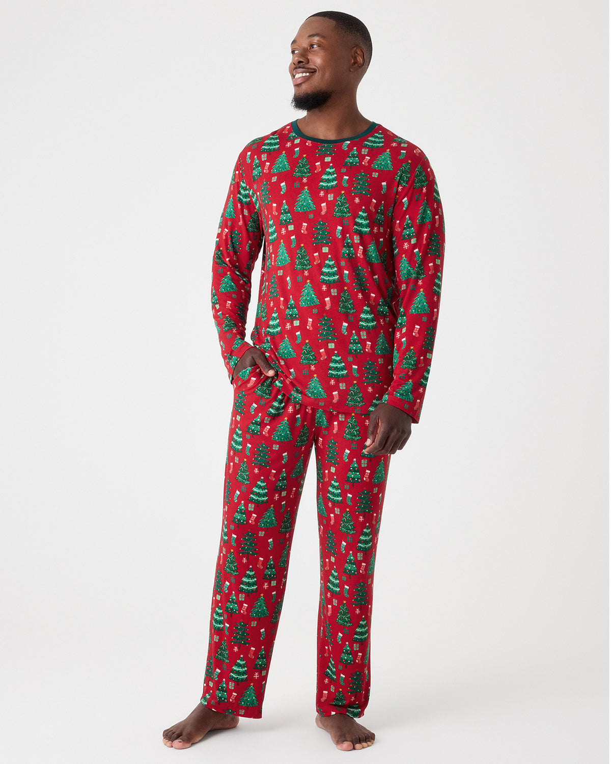 A man wearing a red long-sleeve pajama set with an all-over green Christmas tree pattern, standing against a light gray studio background.