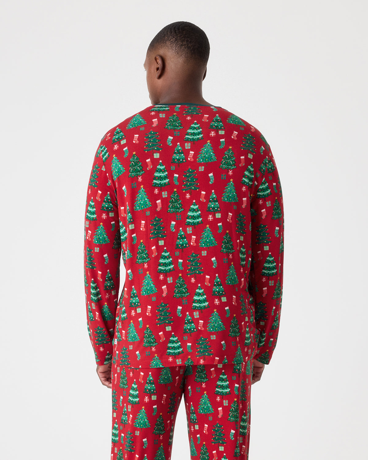 A man viewed from behind wearing a red long-sleeve pajama set with an all-over green Christmas tree and gift pattern, against a light gray studio background.