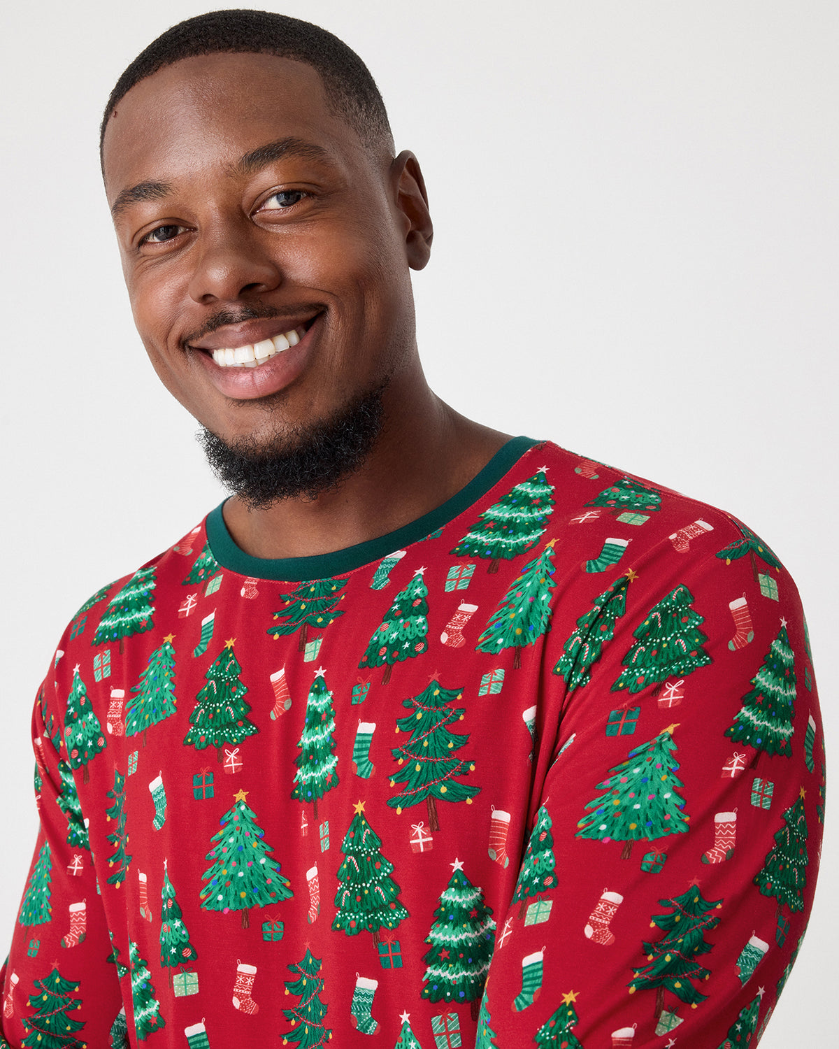A smiling man wearing a red long-sleeve holiday pajama shirt with a green crew neck and an all-over print of green Christmas trees, stockings, and wrapped presents, photographed against a light gray studio background.