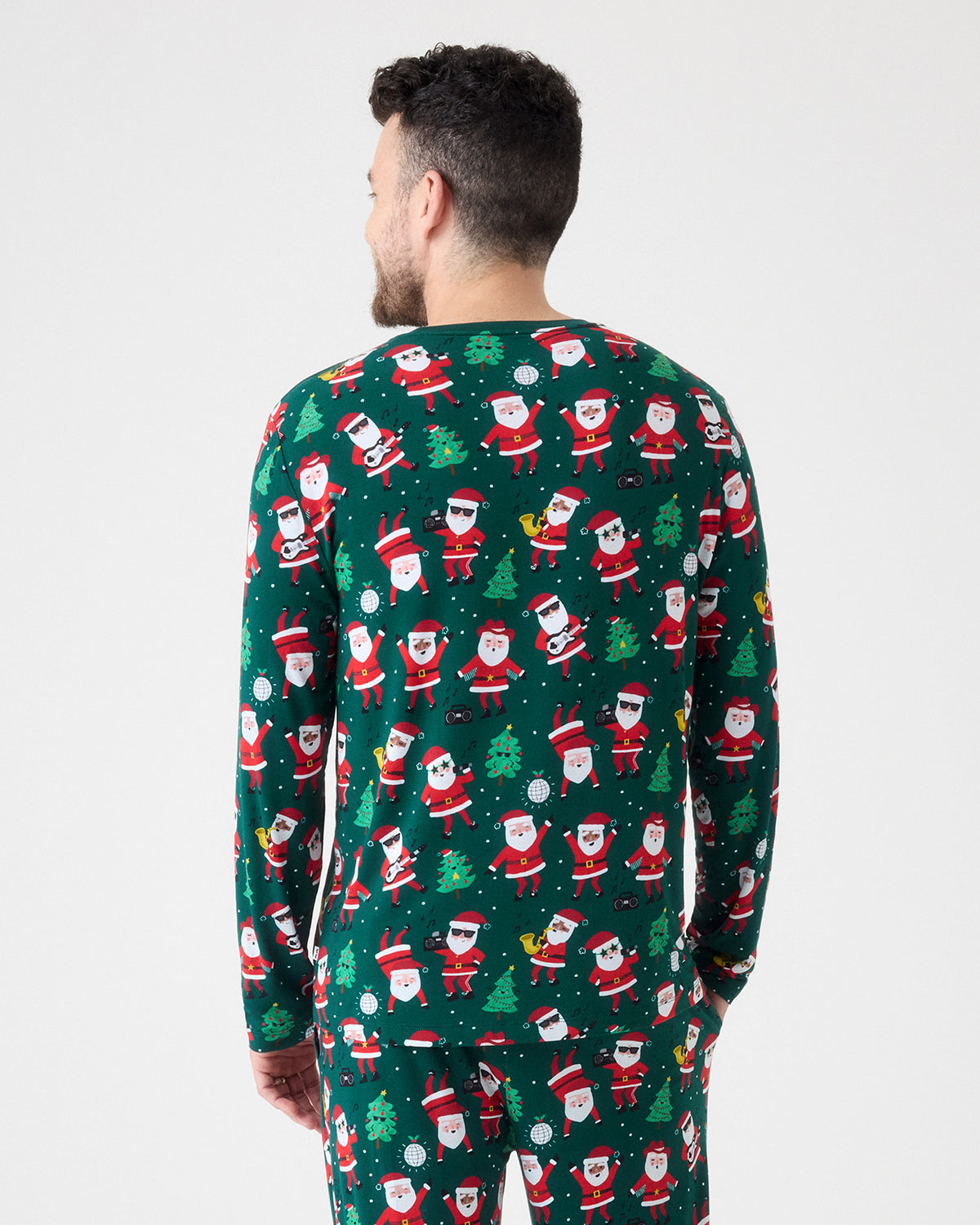 A man viewed from behind wearing a dark green long-sleeve pajama set with an all-over Christmas Santa Claus pattern, against a plain light gray studio background.