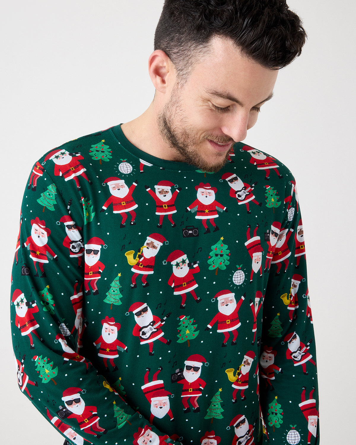 A man wearing a dark green long-sleeve crewneck sweater with an all-over festive print of Santa Claus, Christmas trees, and holiday icons, photographed against a plain light gray background.
