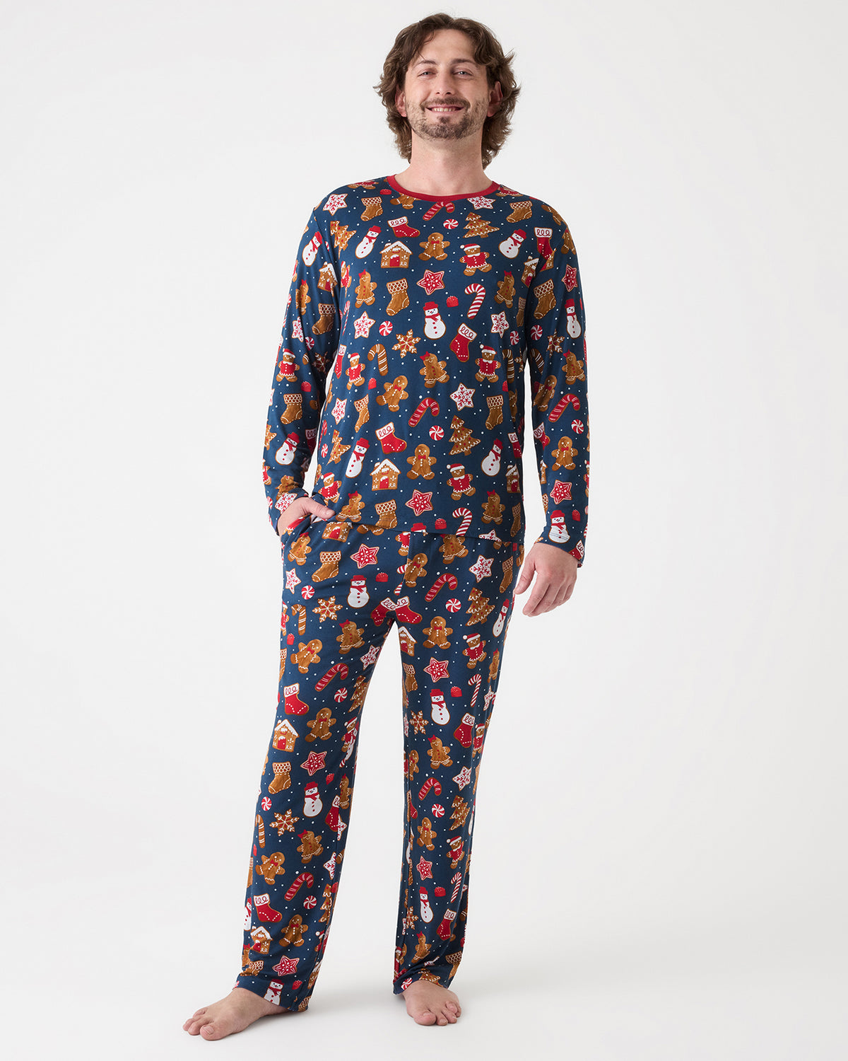 A man wearing a navy blue long-sleeve pajama set with a colorful Christmas-themed print of gingerbread cookies, snowmen, candy canes, and Santa Claus, standing against a plain white studio background.