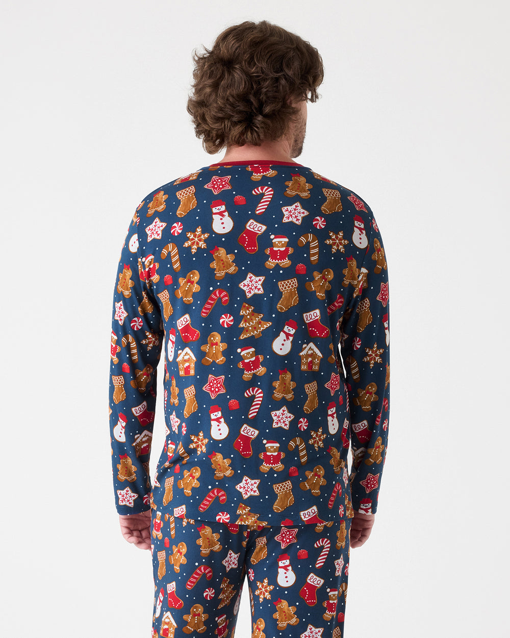 A man viewed from behind wearing a long-sleeve Christmas-themed pajama set with a navy blue background and an all-over festive print of gingerbread men, snowmen, stockings, candy canes, and cookies.